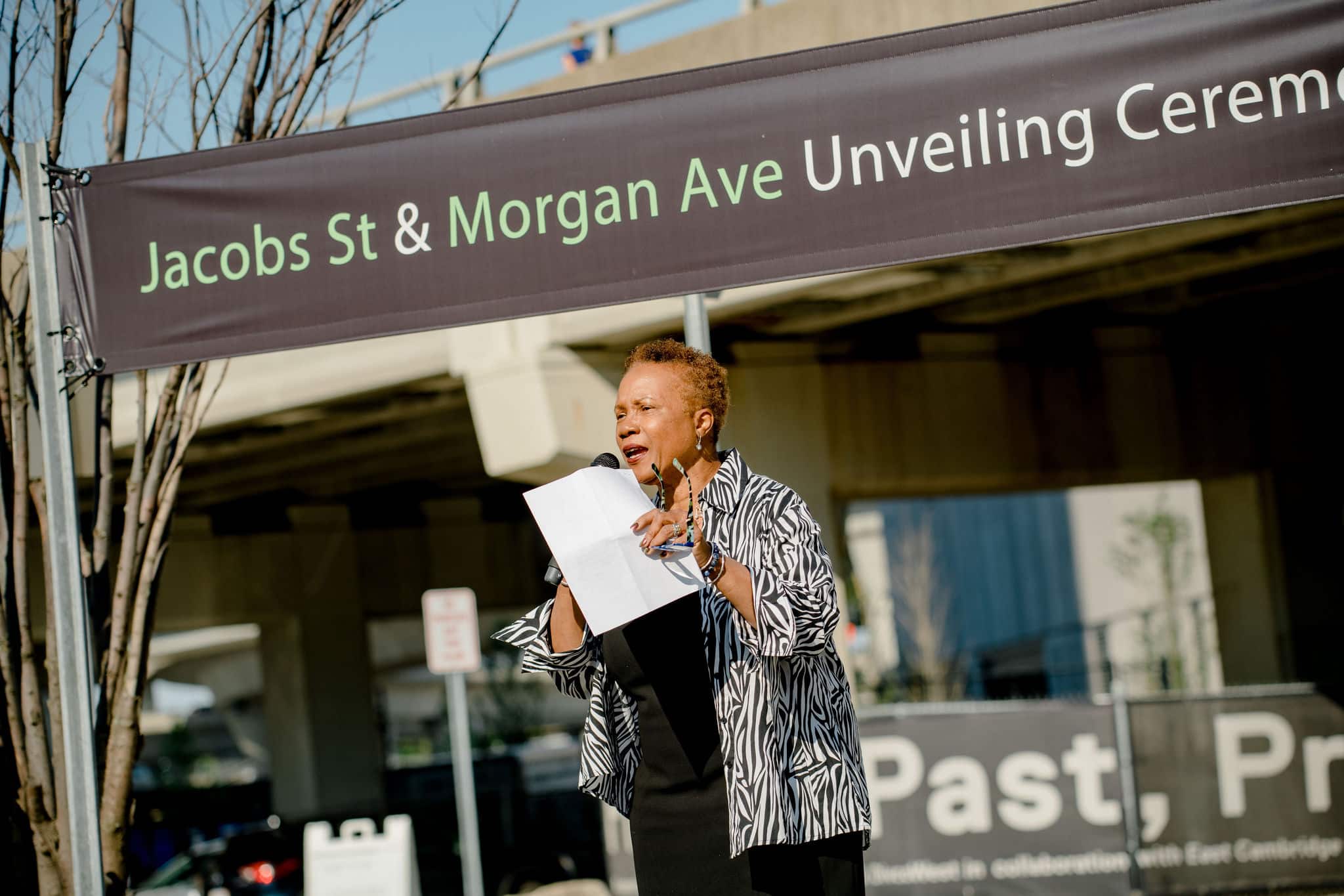 Community leader addresses crowd during street renaming event beneath highway overpass structure