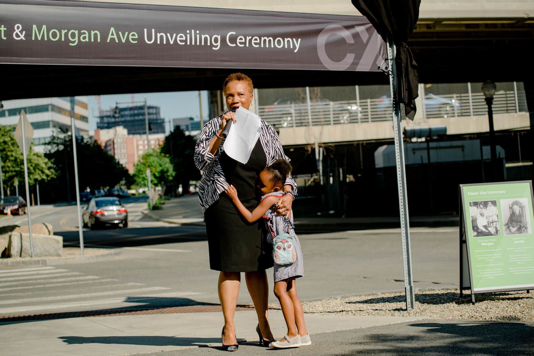 Woman embraces child while speaking at Jacobs and Morgan Ave unveiling ceremony in summer sunlight