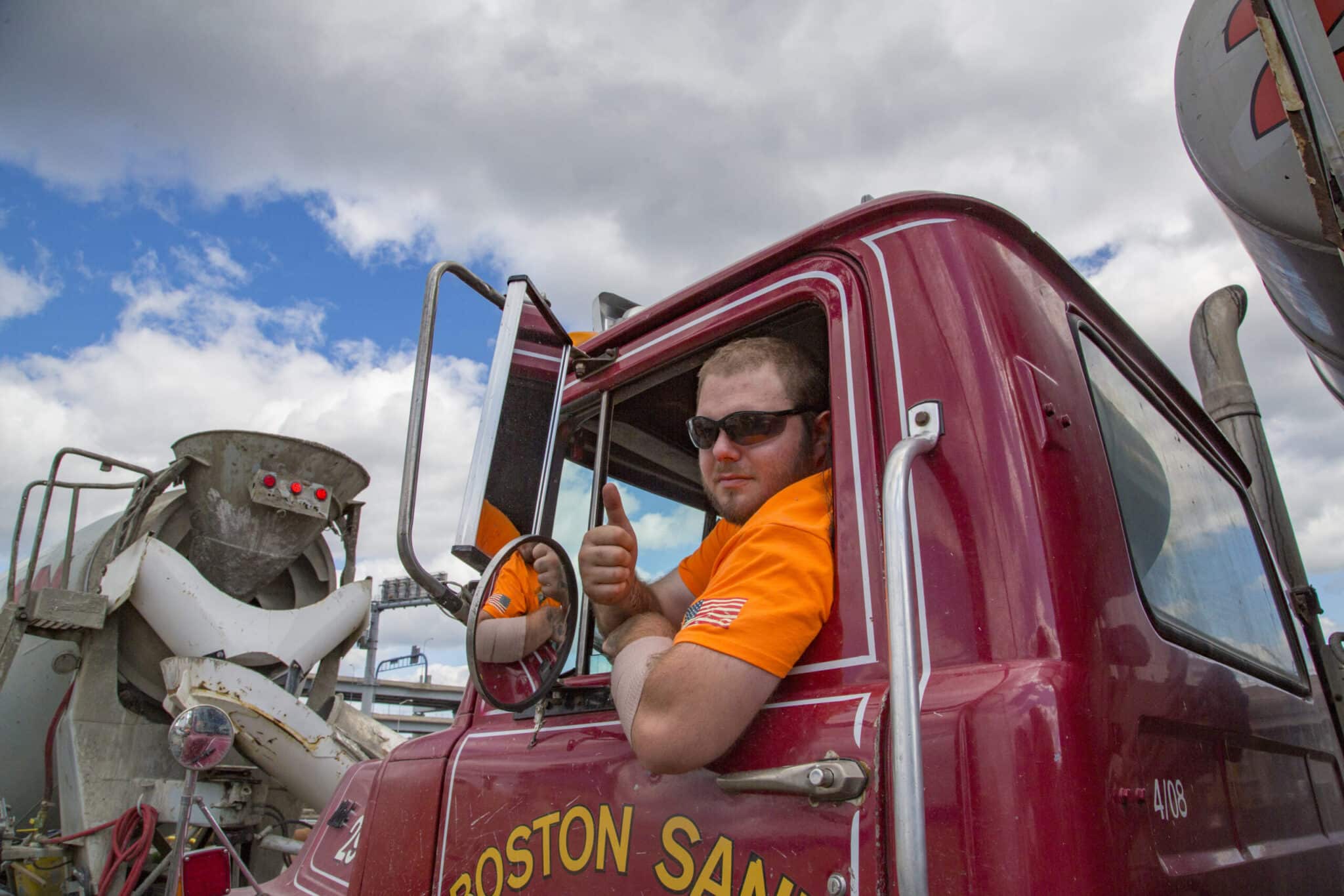 Cement truck driver in orange shirt gives thumbs up from red Boston Sand and Gravel vehicle