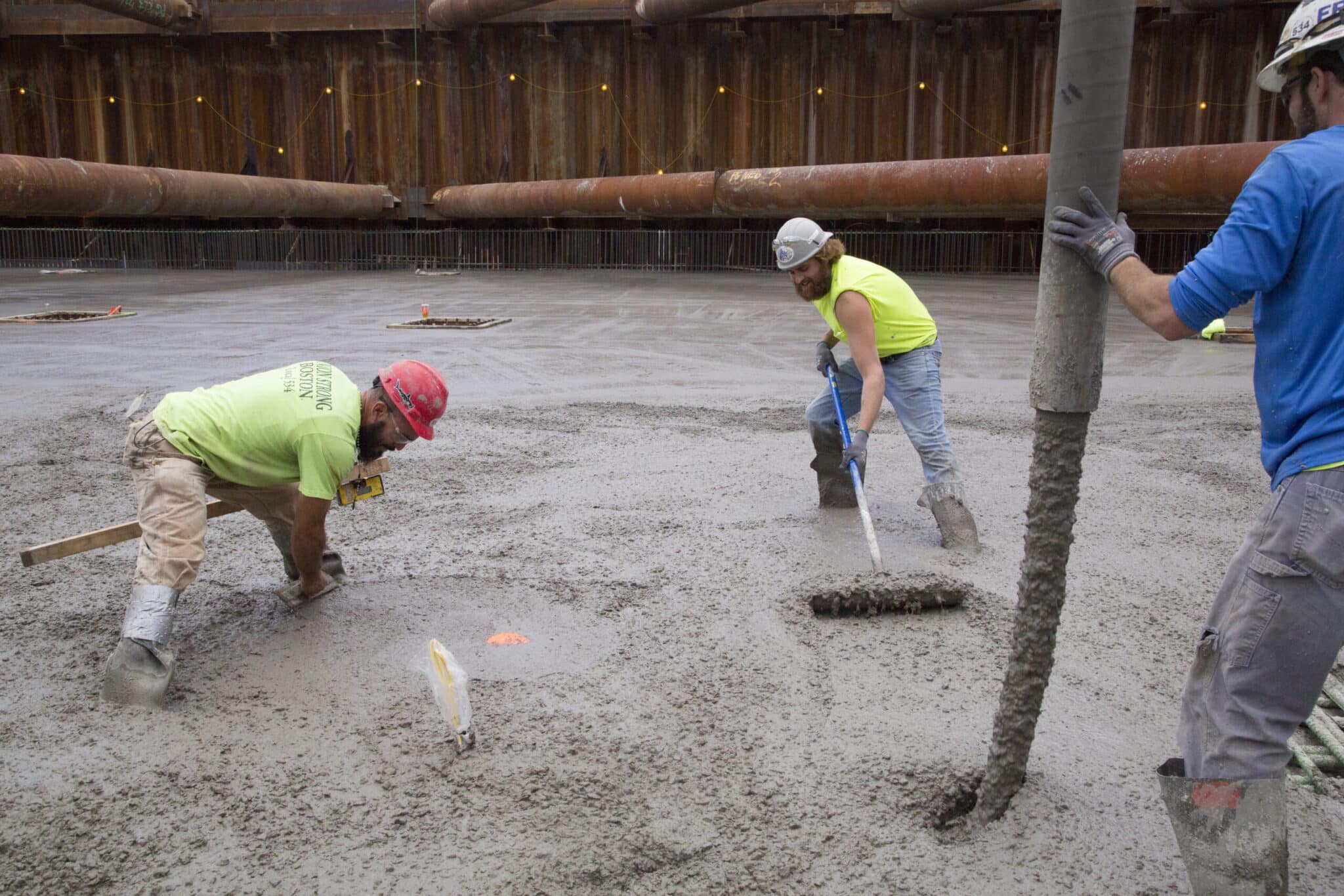 Construction workers smoothing poured concrete foundation with rakes and boots in large pit