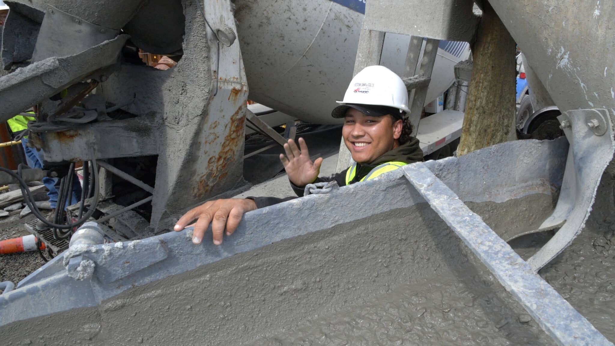 Construction worker in hard hat smiles and waves beside concrete chute at job site