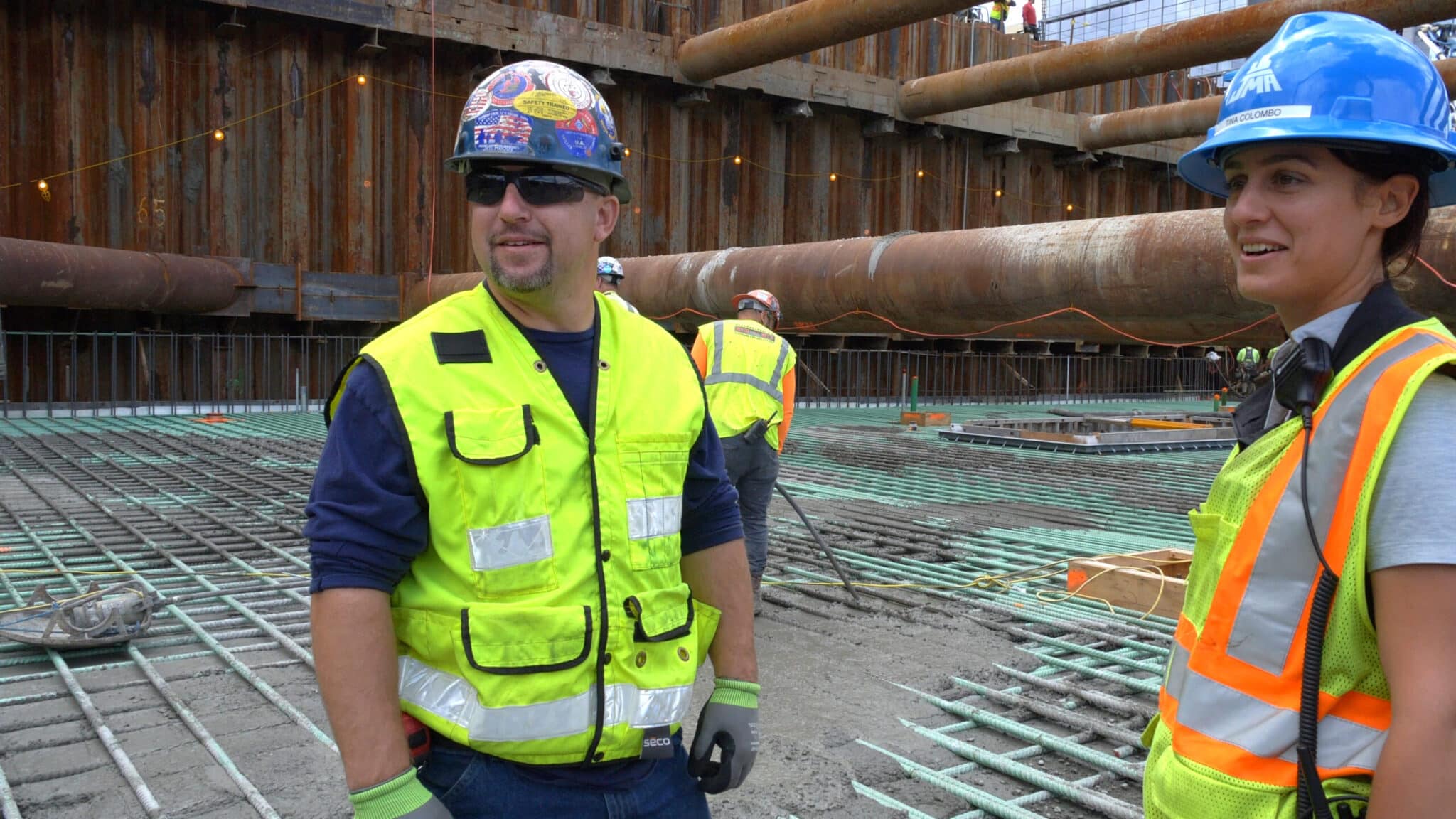 Two workers in safety vests discuss rebar installation on site surrounded by construction materials