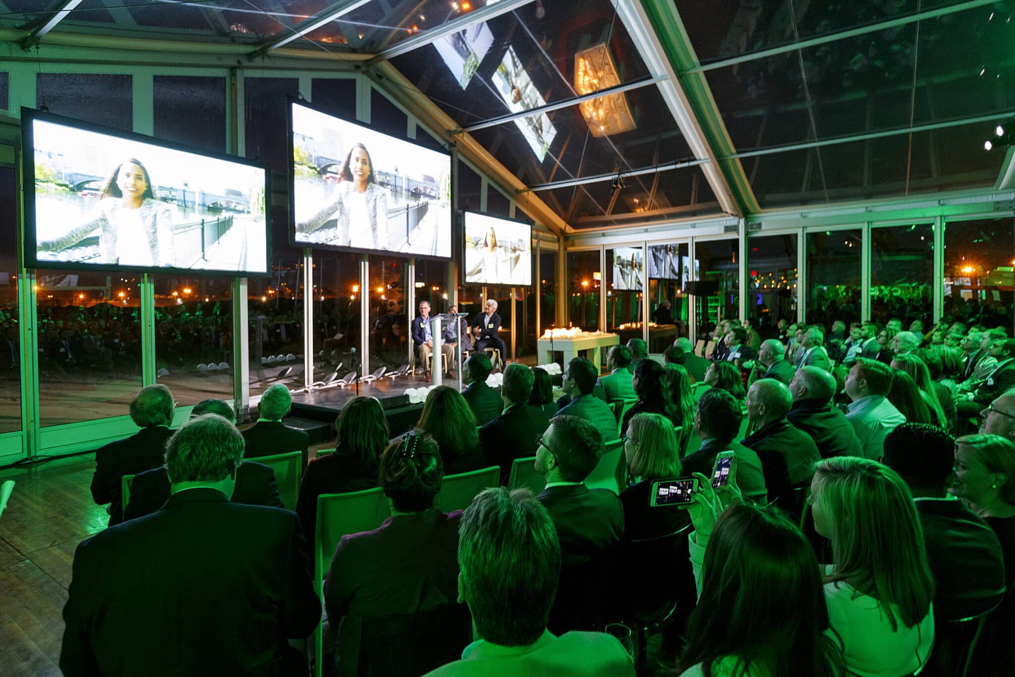 Evening audience watches large screens in a transparent event tent during Cambridge Crossing celebration