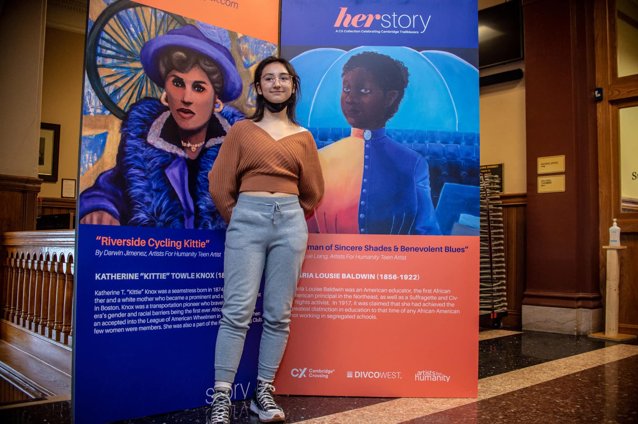 Young woman poses between murals of Katherine Knox and Maria Baldwin at HerStory exhibit