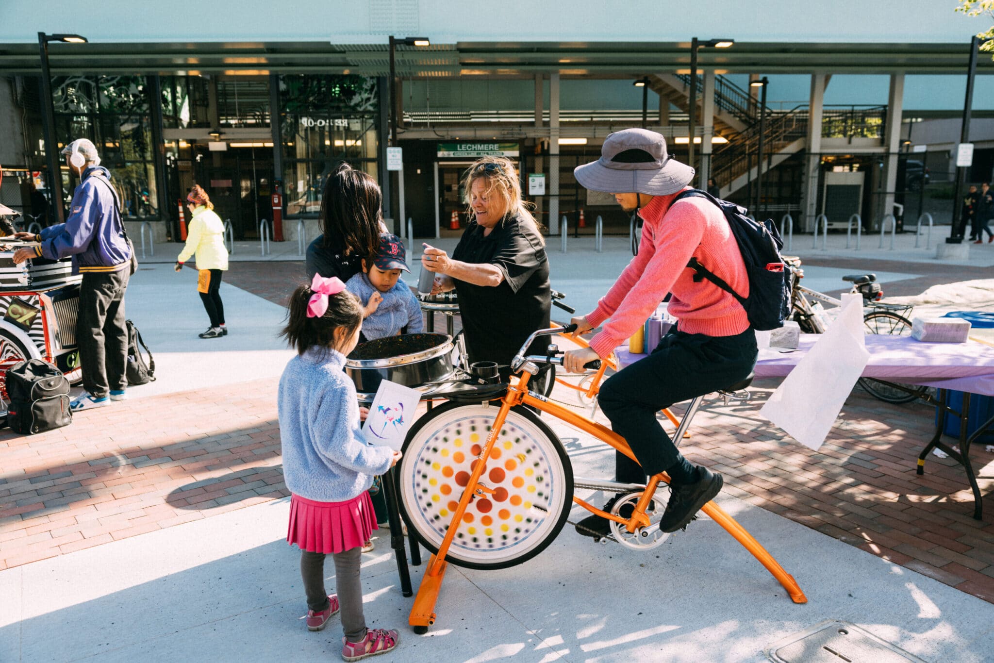Child watches man on orange stationary bike power interactive art spinner at outdoor event