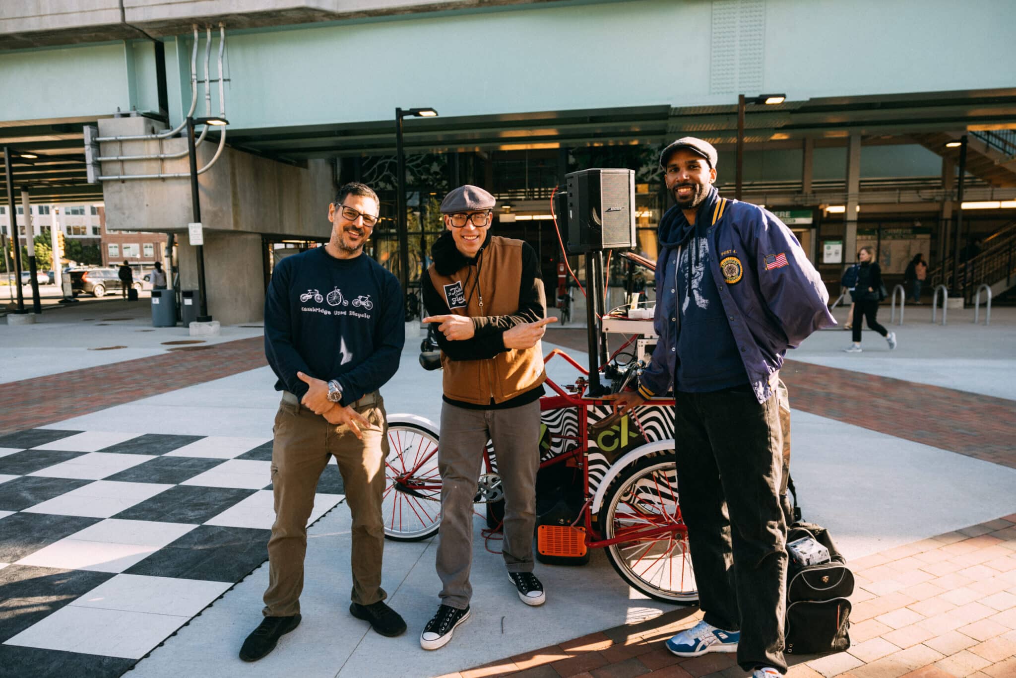 Three men pose beside mobile DJ booth decorated with colorful graphics at outdoor event