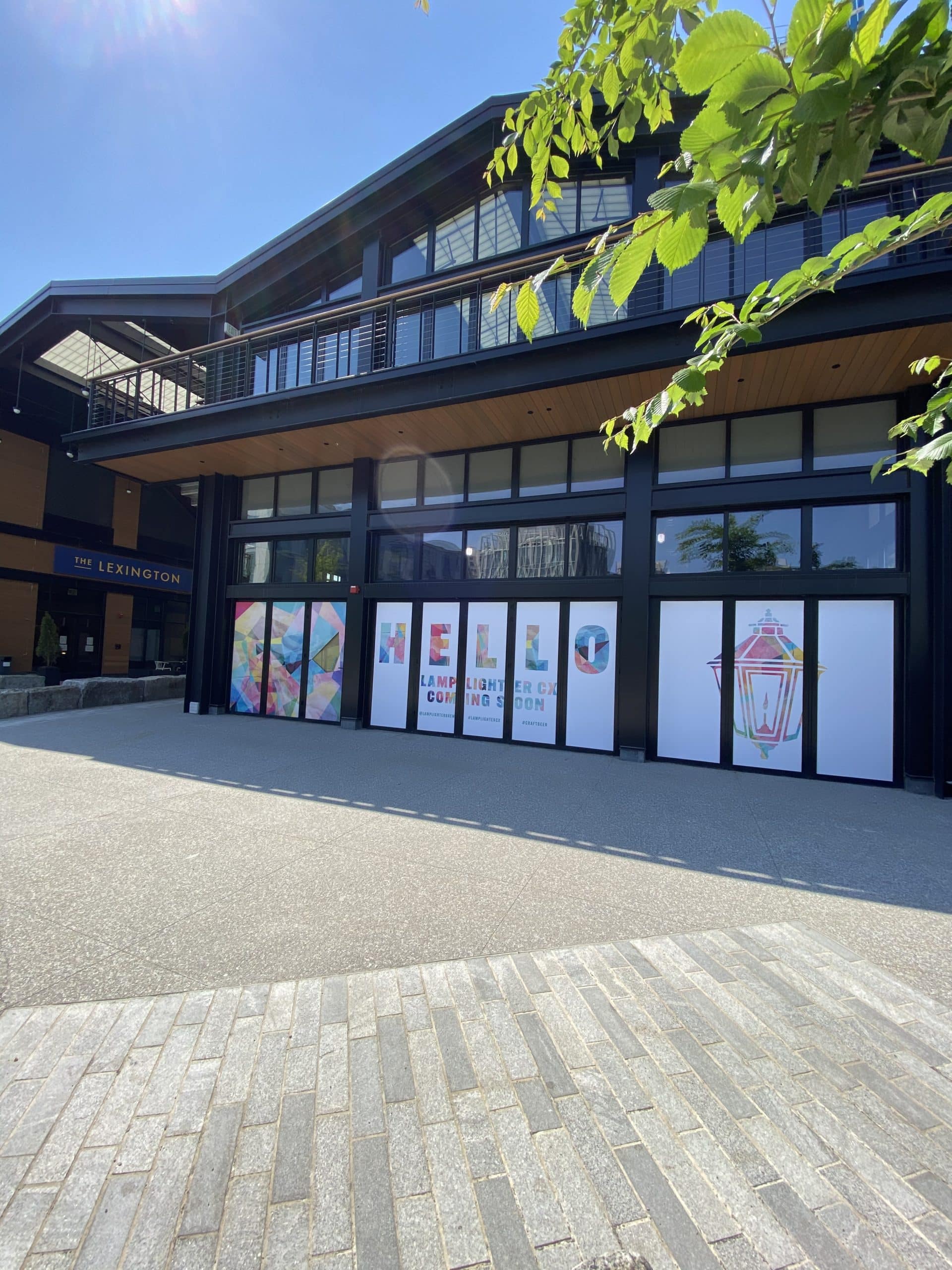 Exterior signage for Lamplighter CX with “Hello” greeting beside neighboring storefronts and plaza