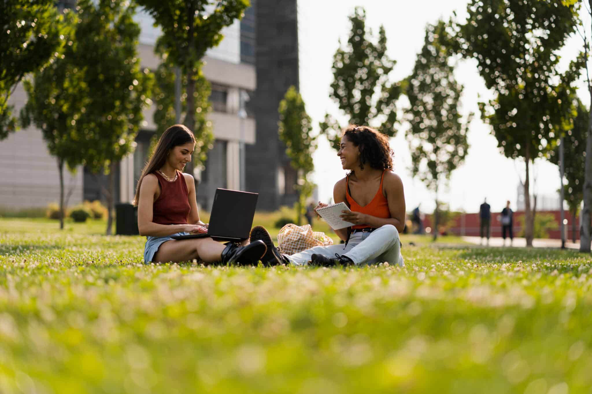 Two women sitting on grass with laptop and notebook near modern buildings and trees