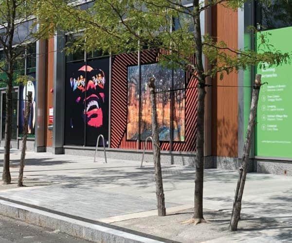 Outdoor urban mural with vibrant pop-art face and colorful landscape displayed behind tree row