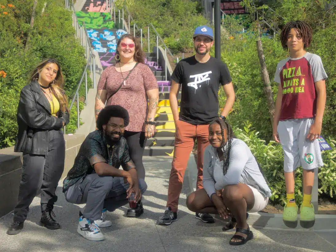 Group of six people posing proudly in front of colorful outdoor staircase mural installation