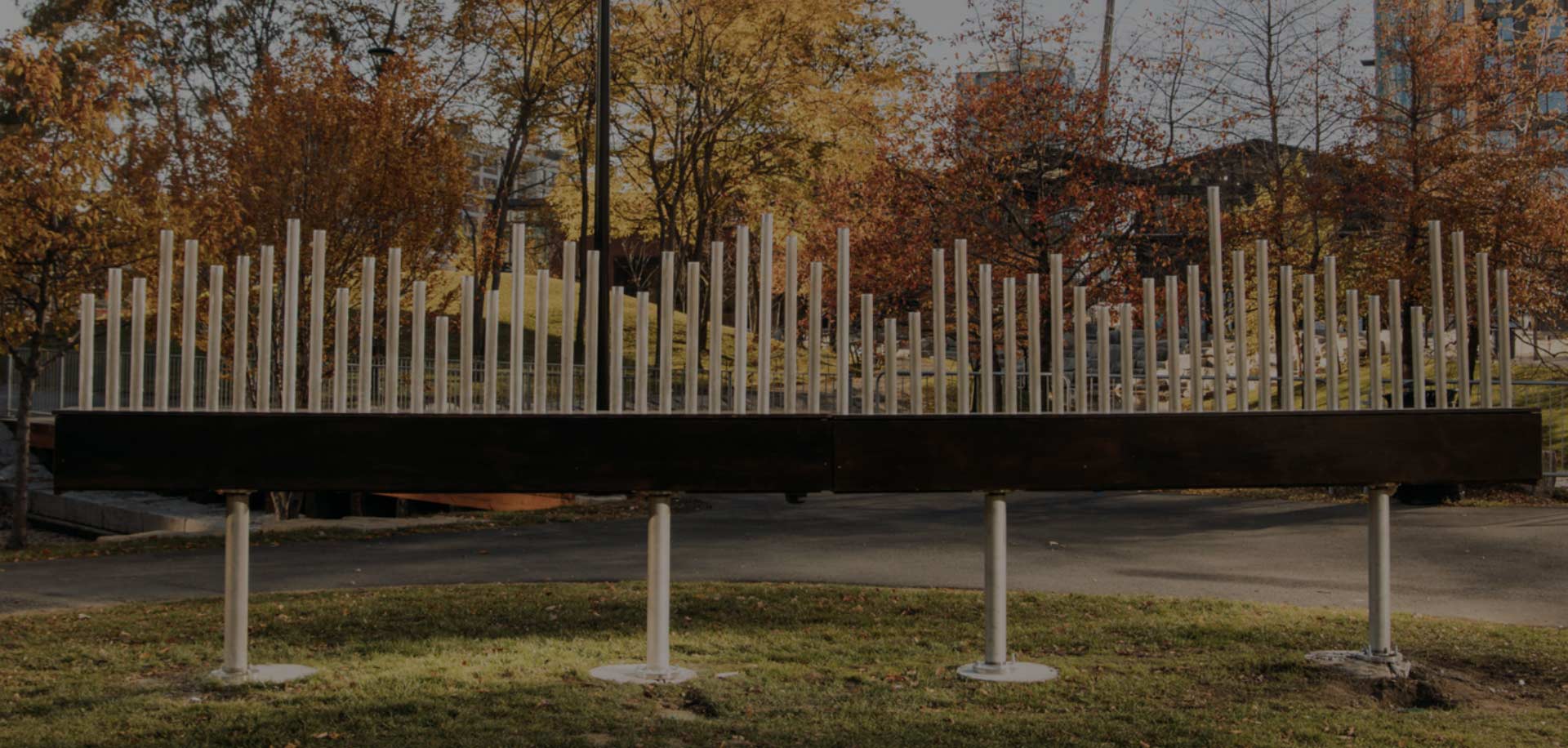 Urban park sculpture featuring vertical metal pipes on a black base, surrounded by fall foliage