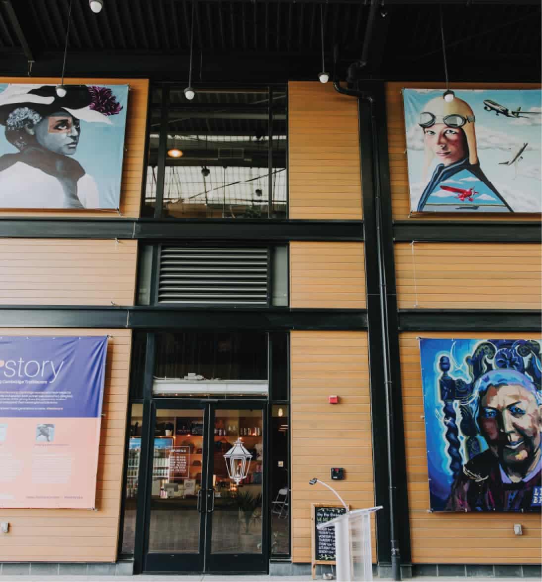 Public indoor mural display of three diverse women portraits above storefront entrance in bright gallery