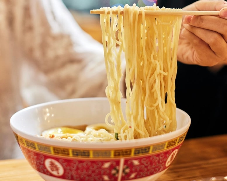 Close-up of hand lifting fresh ramen noodles with chopsticks from decorated Asian-style bowl