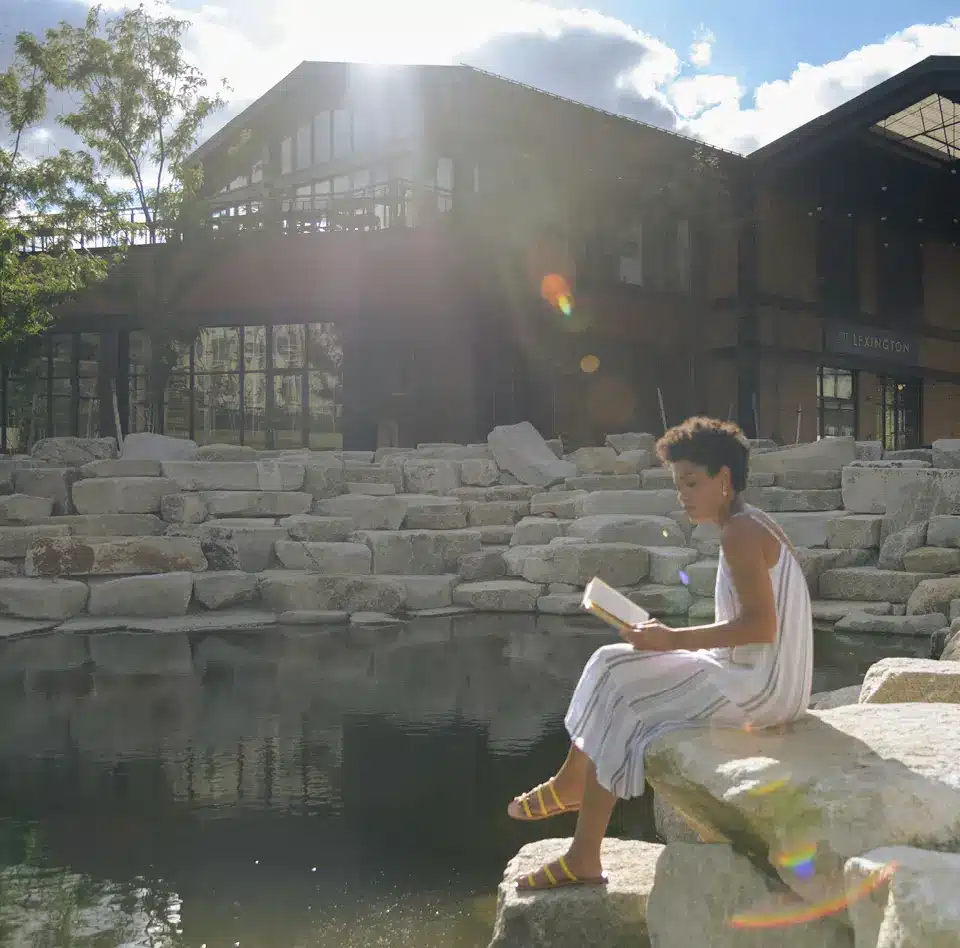 Woman reading by stone pond steps at Lexington restaurant, sun flare beaming across tranquil water