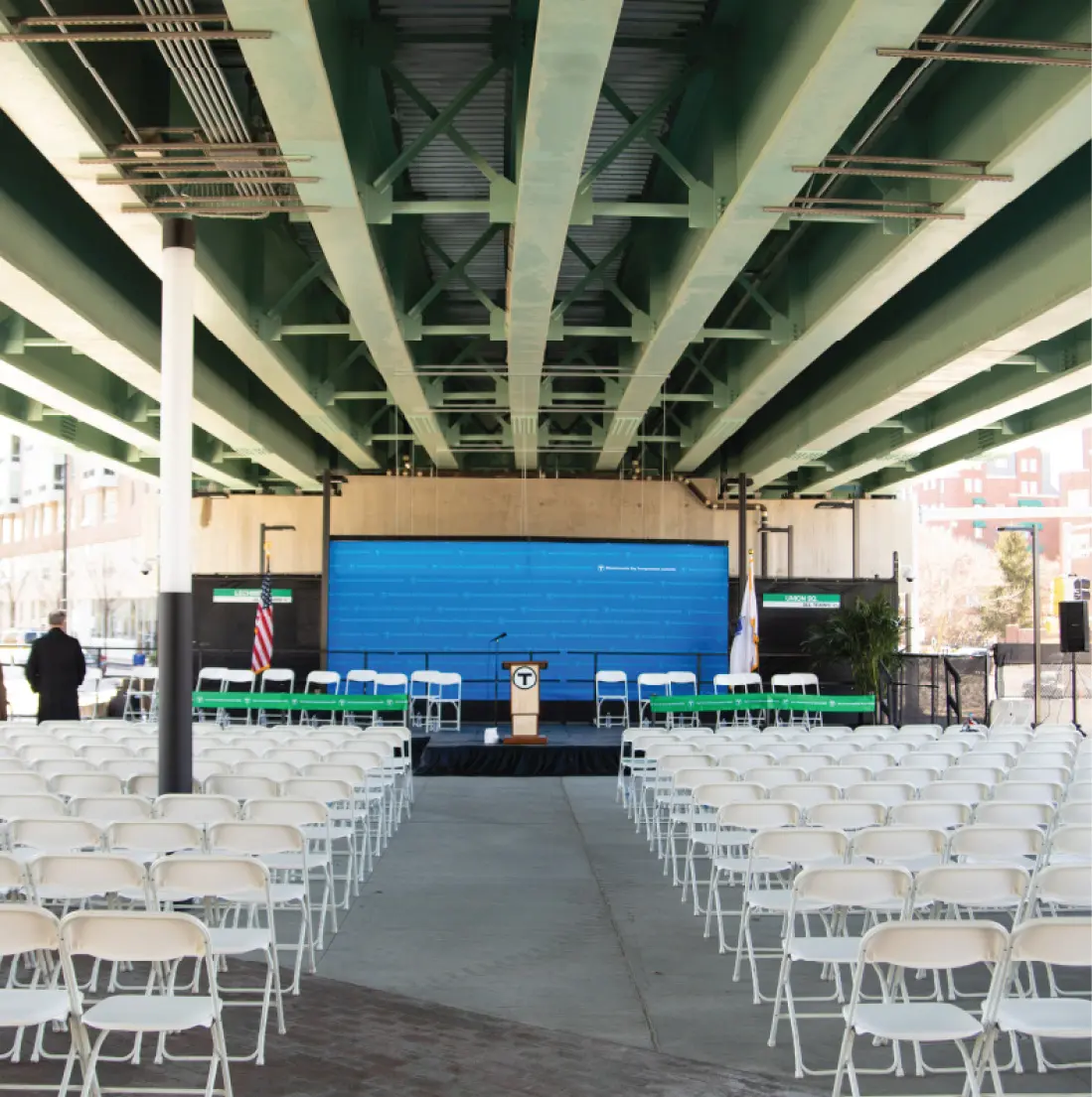 Outdoor stage setup beneath highway bridge with rows of empty white chairs and blue backdrop