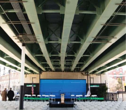 Wide-angle view of urban stage setup under green metal bridge with chairs aligned in rows
