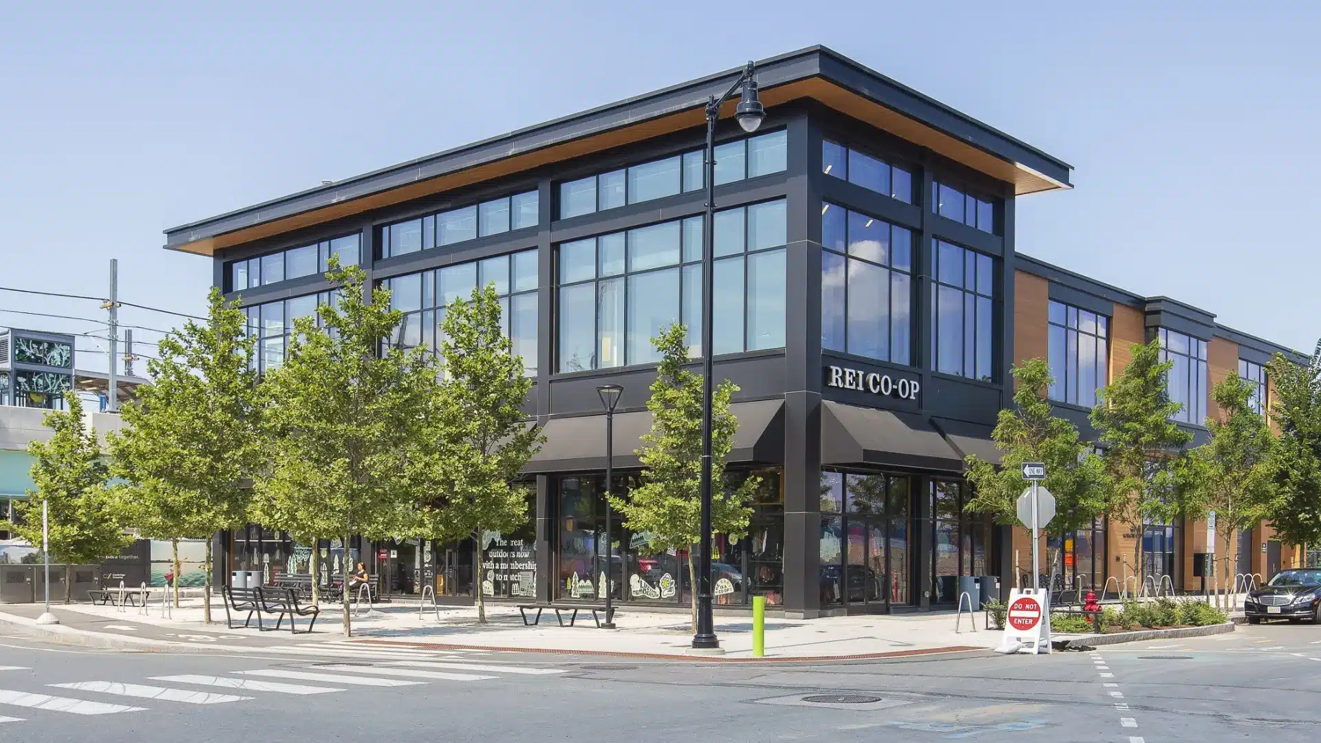 Modern corner retail building with REI Co-op signage surrounded by trees and street furniture