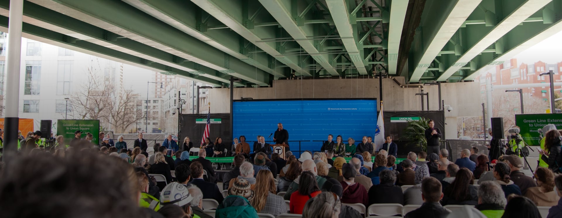 Press conference under highway overpass featuring Massachusetts Bay Transportation Authority signage