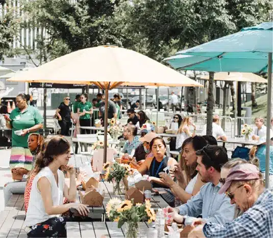 Crowded outdoor seating area with people sharing food under colorful sun umbrellas