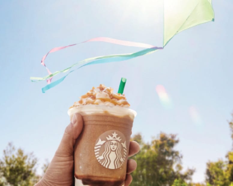 Hand holding Starbucks caramel Frappuccino drink with a colorful kite flying above