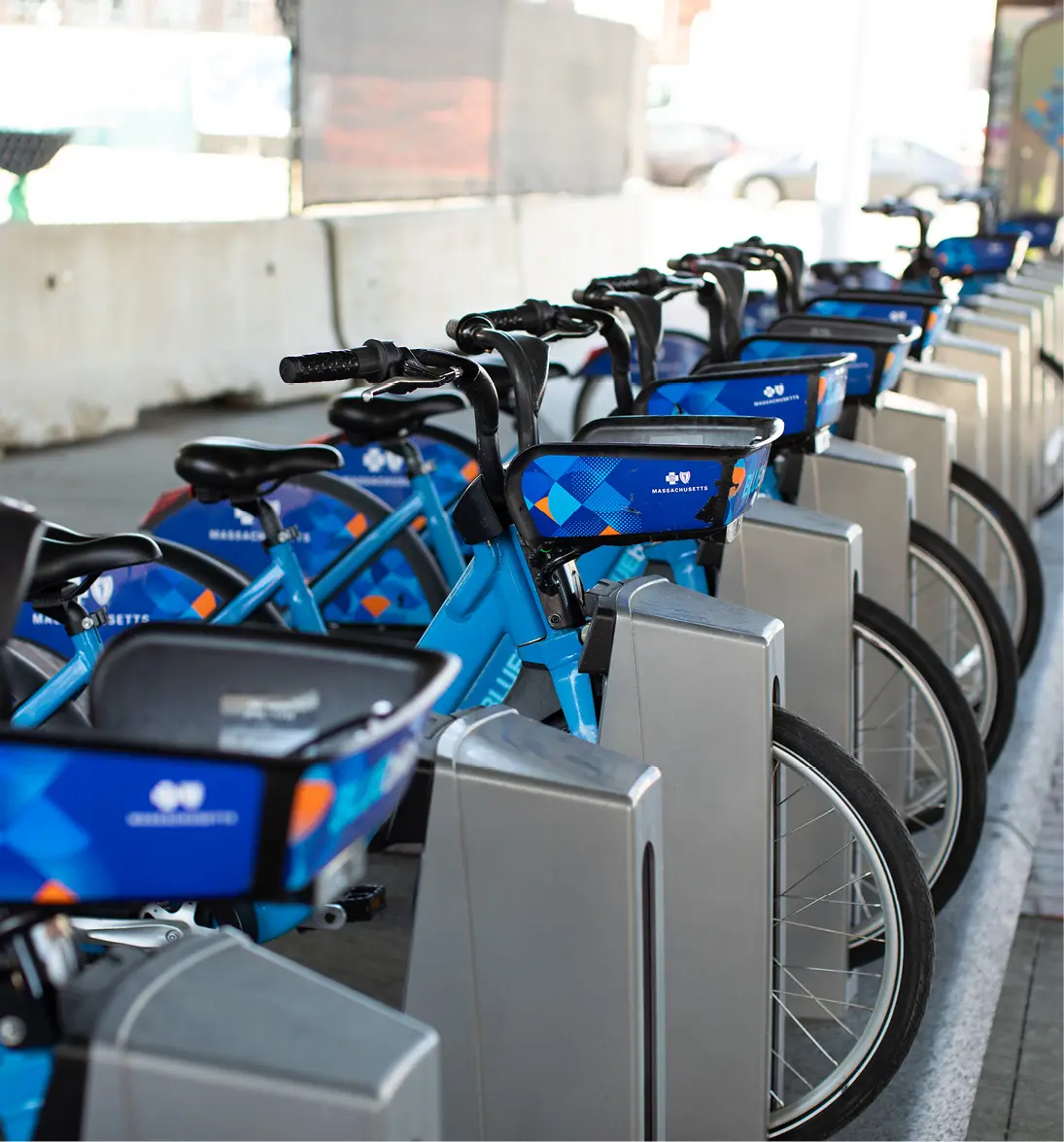 Row of blue Massachusetts Bluebikes parked in urban bike-share station near concrete barrier