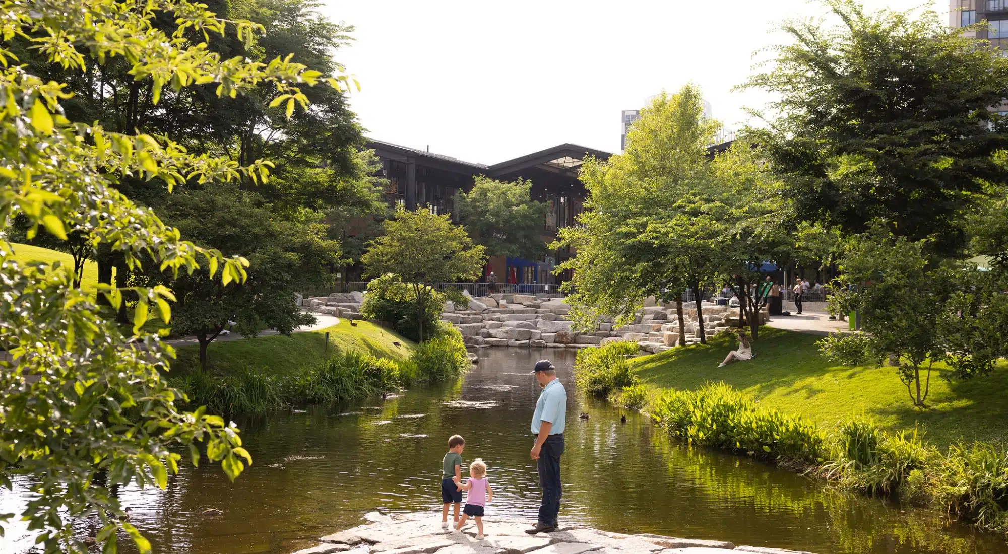 Serene landscaped public space with stepping stones, trees, and small children near gentle stream